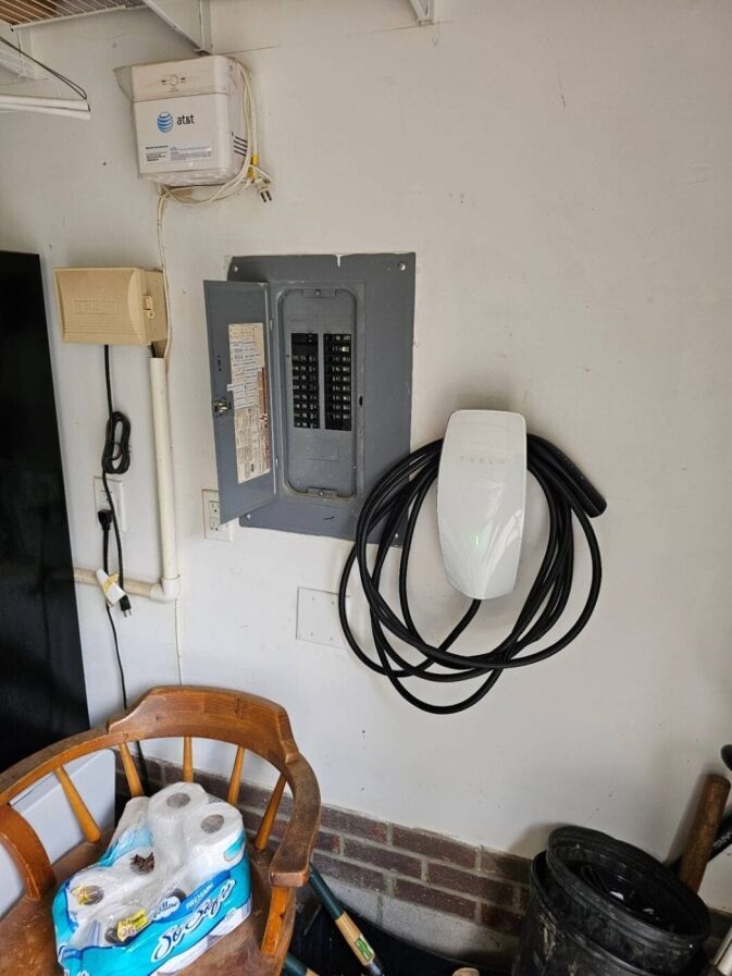 An electrical panel with its door open, a Tesla wall charger mounted beside it, and a wooden chair with paper towels in a garage.