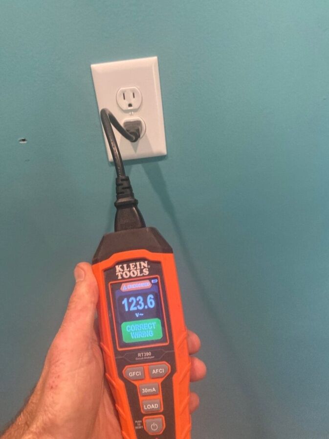 A hand holds a Klein Tools electrical tester showing 123.6V and Correct Wiring while plugged into a wall outlet on a teal-colored wall.