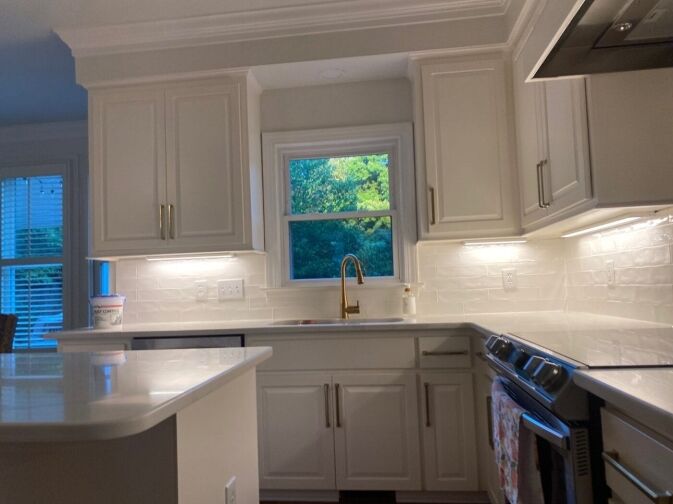 Modern kitchen with white cabinets, under-cabinet lighting, a gold faucet, stainless steel appliances, and a window above the sink showing green trees outside.