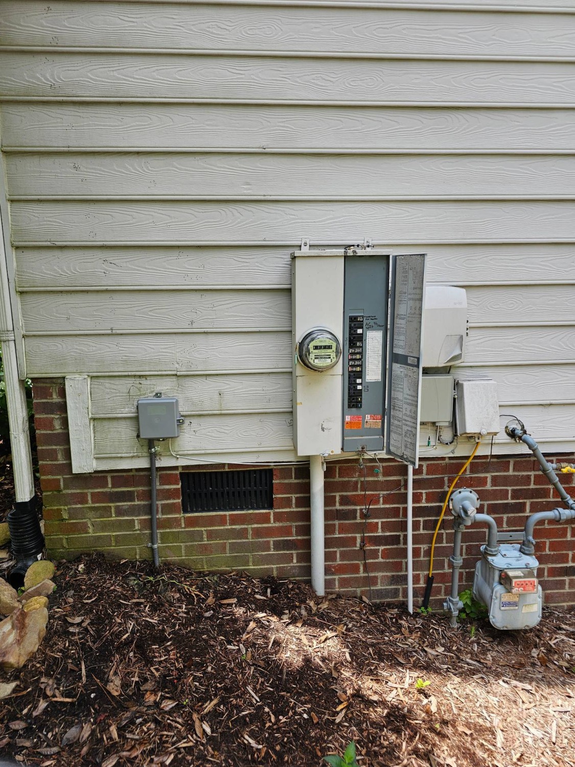 An exterior view of a house wall showing an electric meter, an open electrical panel, conduit, and a gas meter, with mulch and soil in the foreground.