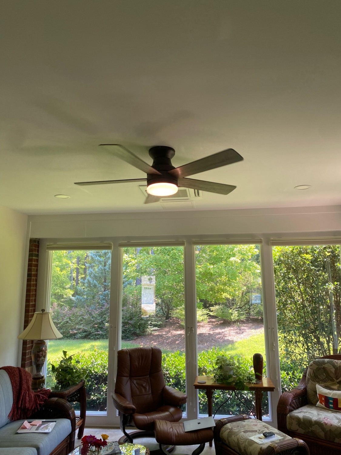 A living room with large windows, green outdoor view, a ceiling fan with light, two armchairs, and a side table with magazines.