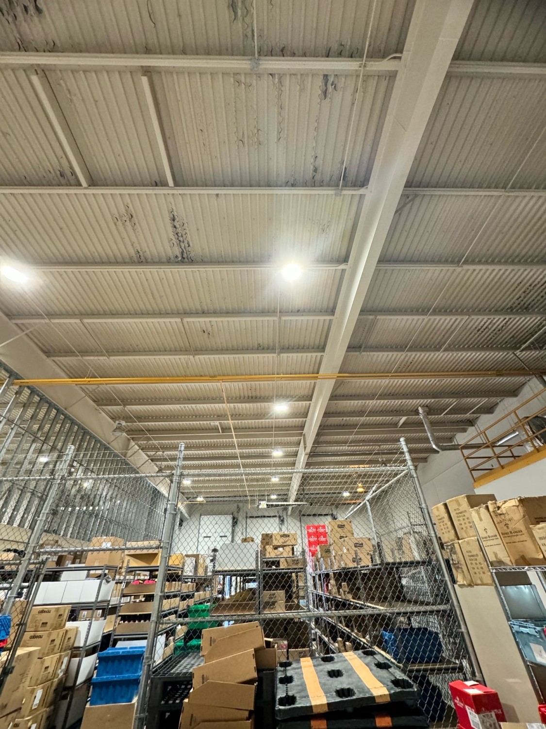 Warehouse interior with metal shelving, cardboard boxes, and bins stacked; fenced area in foreground and exposed ceiling with lights above.