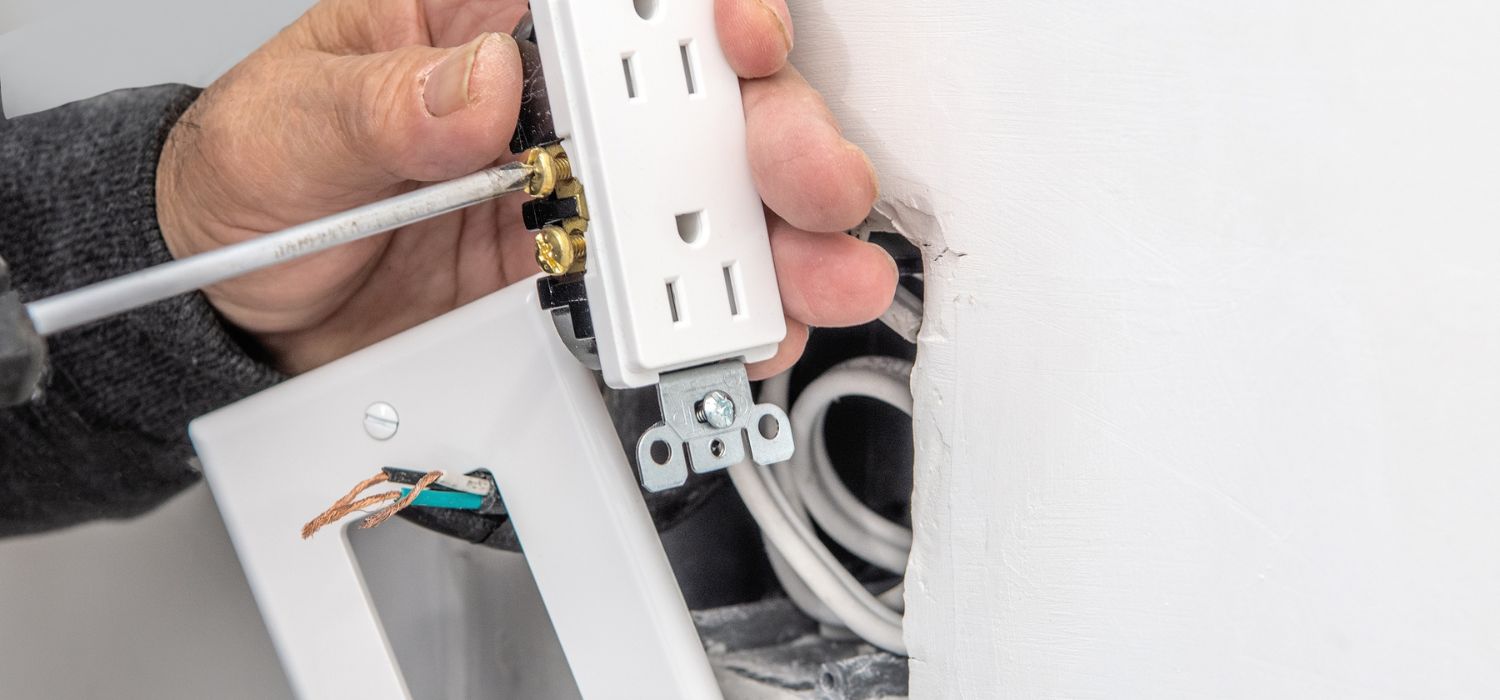 A person installs an electrical outlet into a wall using a screwdriver, with visible wires and an outlet cover nearby.