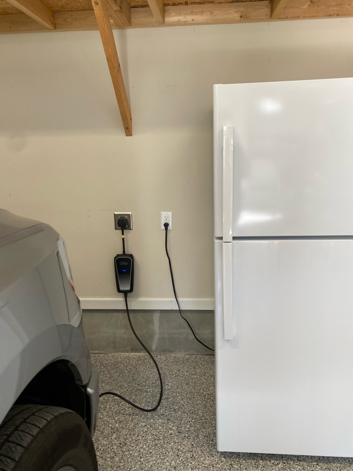 A gray car parked in a garage next to a white refrigerator and a wall-mounted electric vehicle charger plugged into a power outlet.