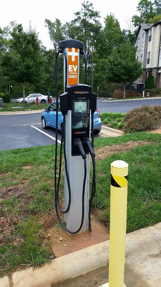 Electric vehicle charging station in a parking lot with two charging cables and a sign reading EV Only. A blue car is parked nearby. Trees and an apartment building are in the background.
