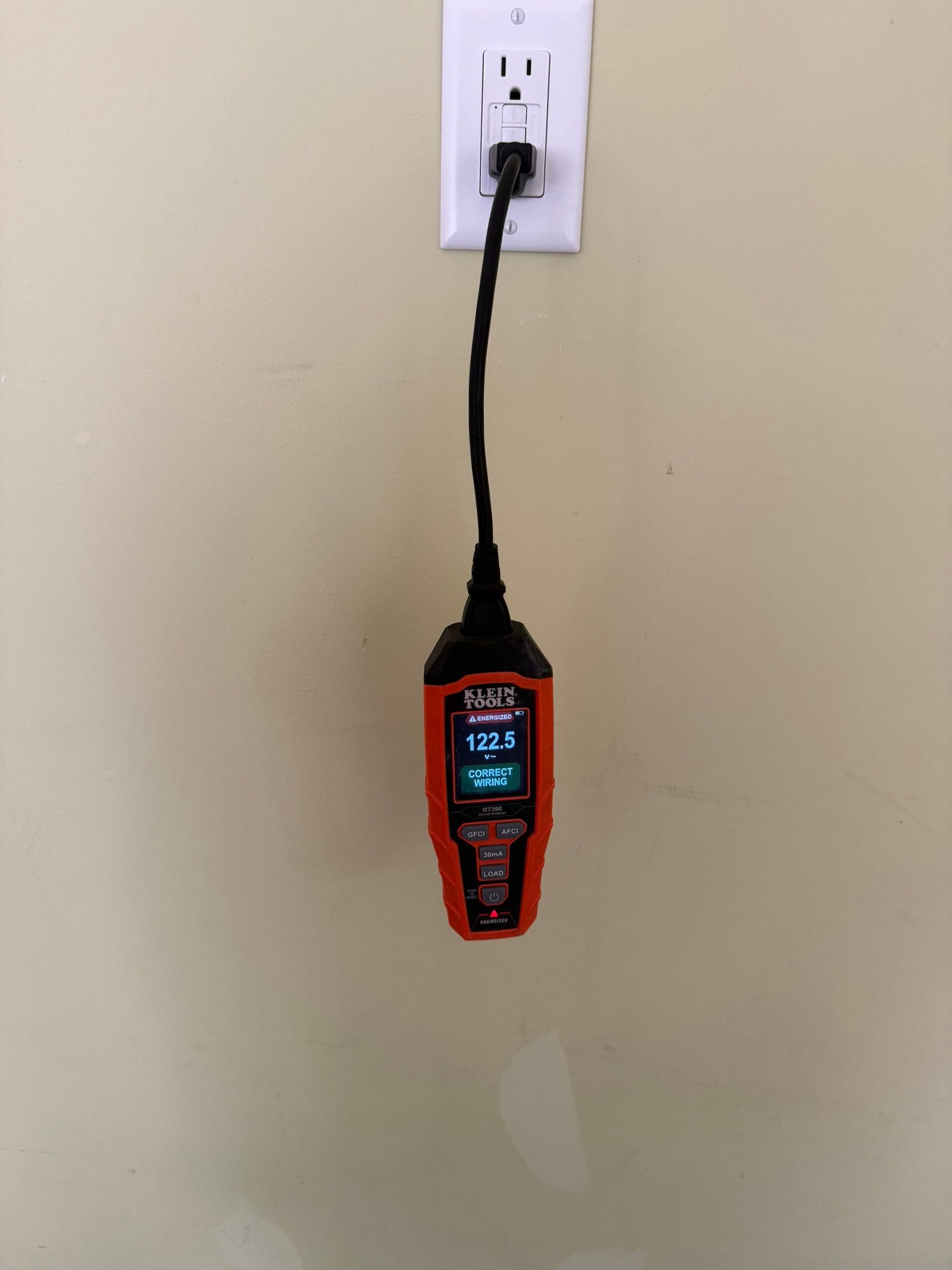A digital voltage tester is plugged into a wall outlet, displaying a reading of 122.5 volts on its screen.