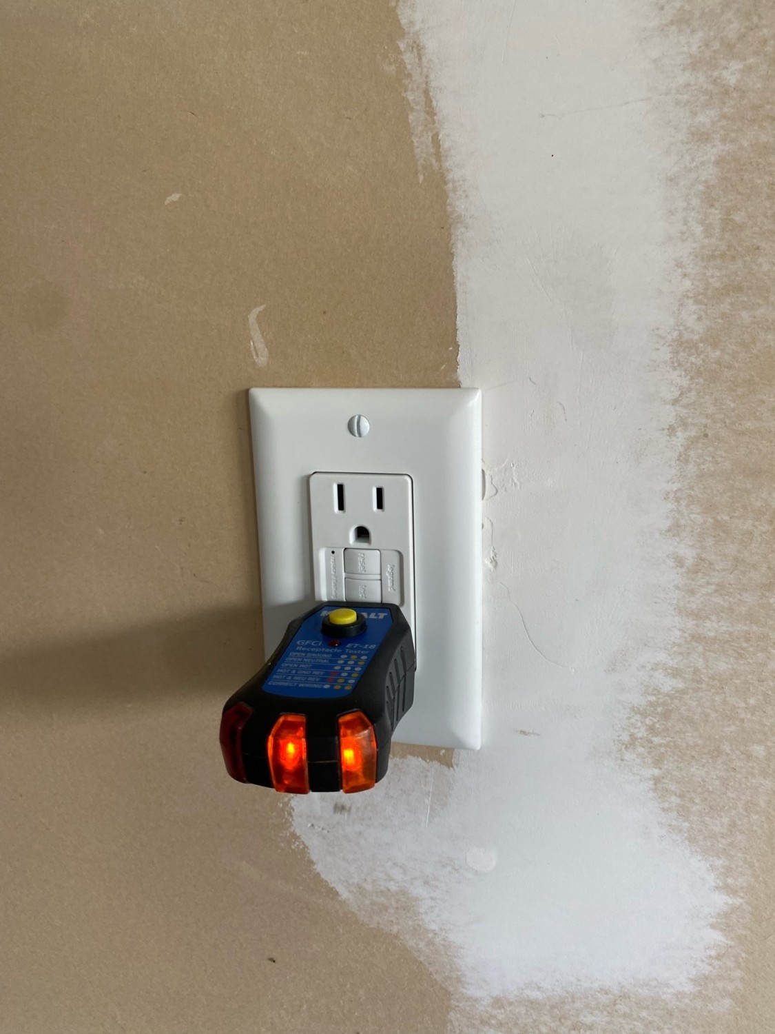 A GFCI electrical outlet with a tester plugged in; two orange lights on the tester are illuminated. The wall shows unpainted drywall patching.