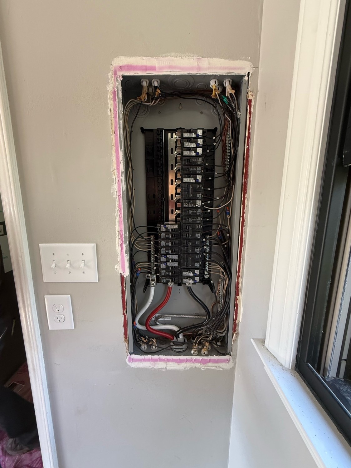 An open electrical breaker panel with multiple circuit breakers and wires is mounted in a wall next to a window and a light switch.