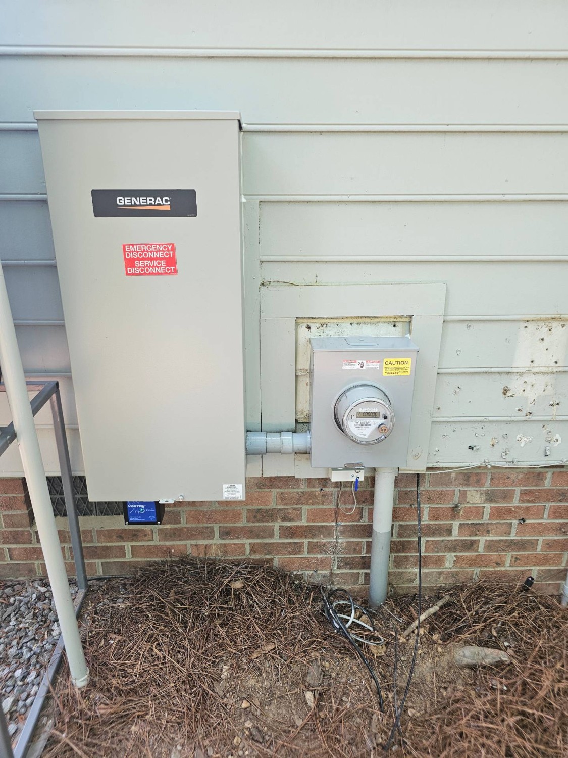A Generac electrical panel and a utility meter are mounted on the outside wall of a house above a brick foundation.