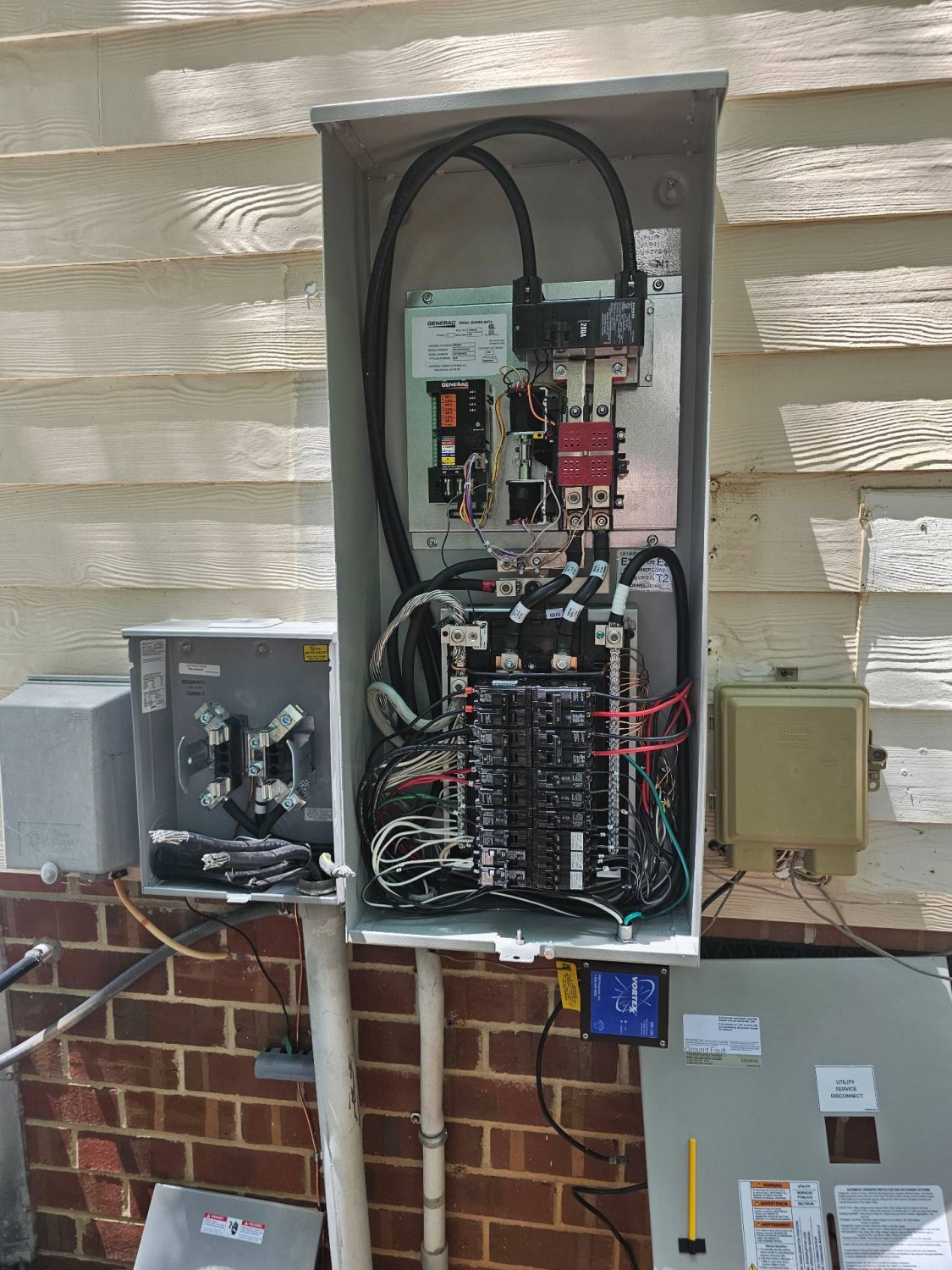 An open electrical panel mounted on an exterior wall reveals circuit breakers, wiring, and connected conduits. Other utility boxes are nearby.