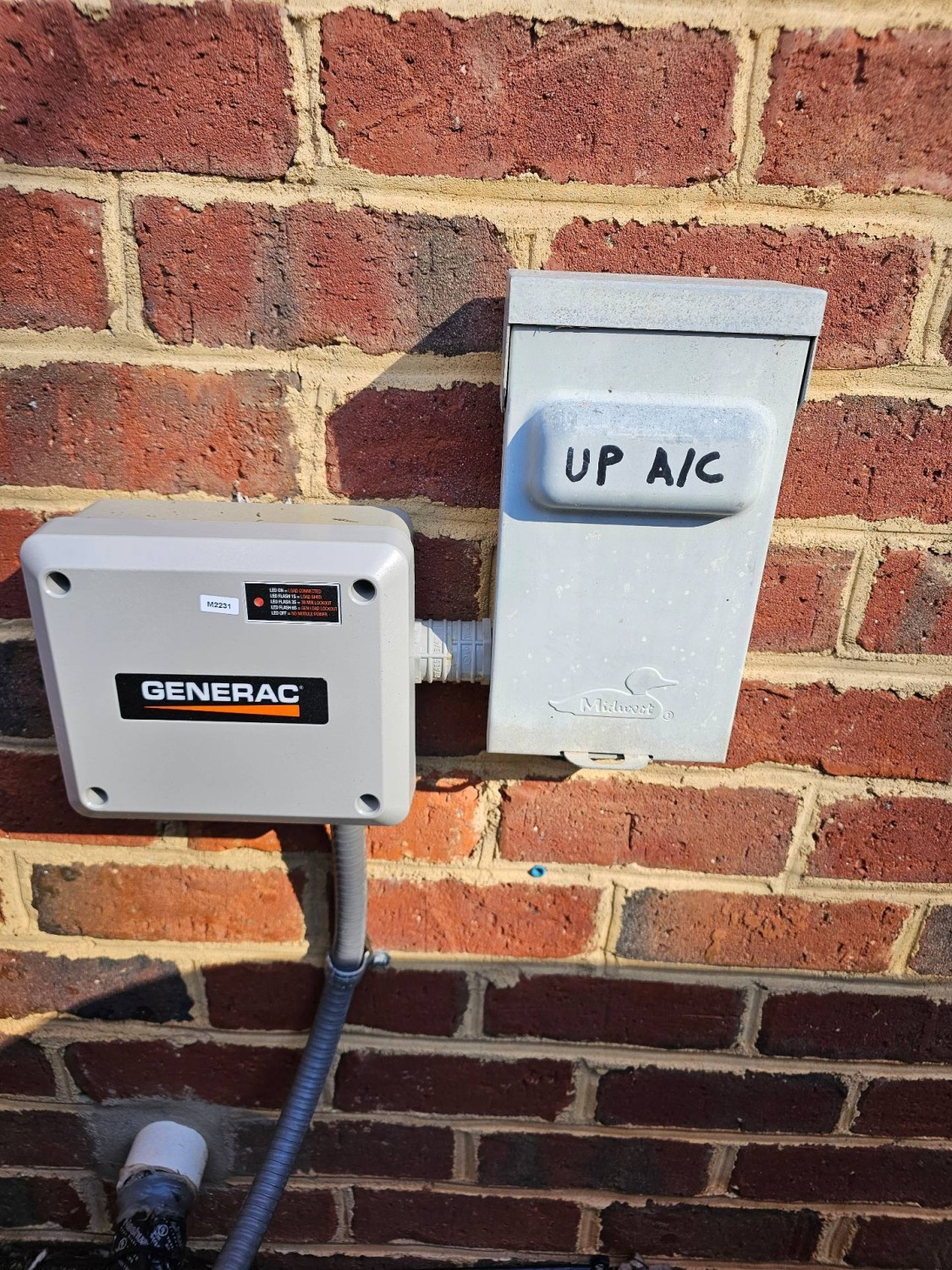 A Generac electrical box and a metal cover labeled UP A/C are mounted on a brick wall. Conduits run into both boxes from below.