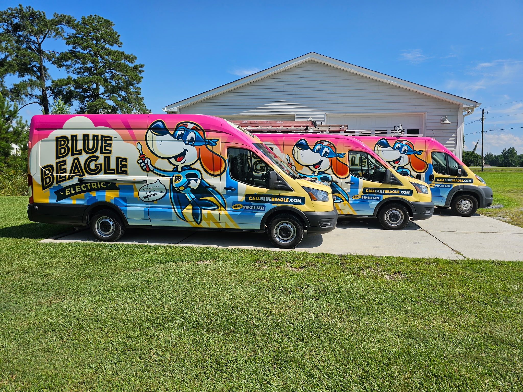 Three colorful Blue Beagle Electric service vans, featuring cartoon dog graphics, are parked side by side on a driveway in front of a white building, ready for their next electrician service call.