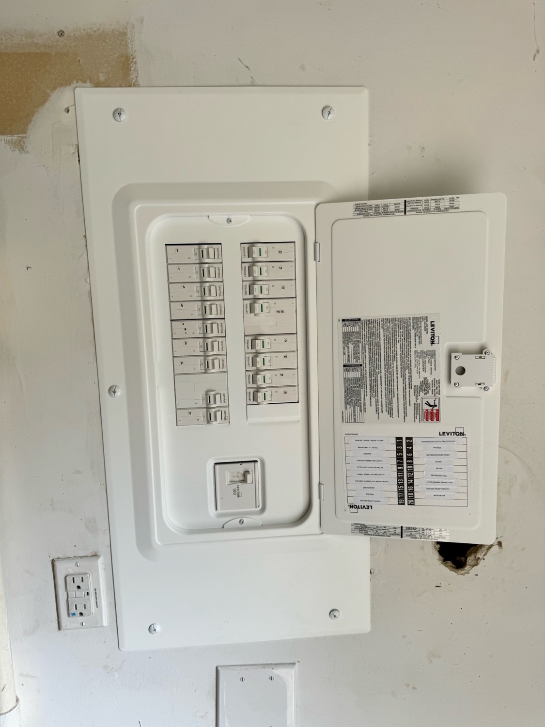 A white electrical breaker panel with its door open, showing labeled circuit breakers and a chart; outlets and wall markings are visible nearby.