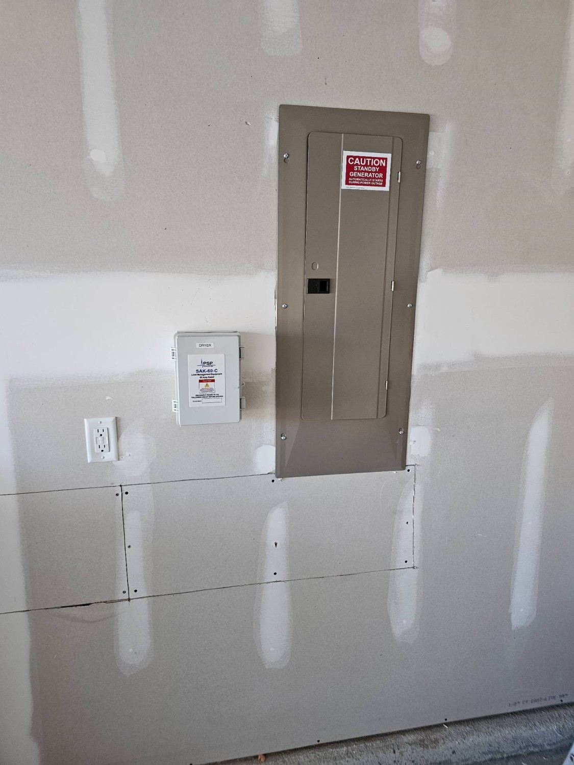 A metal electrical panel and a smaller white utility box are mounted on an unfinished drywall wall, next to a power outlet. Both boxes have caution labels.