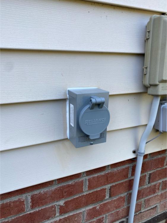 A gray Reliance electrical box, likely installed by an electrician, is mounted on the exterior siding of a house above a brick foundation, next to a utility conduit and another beige utility box.